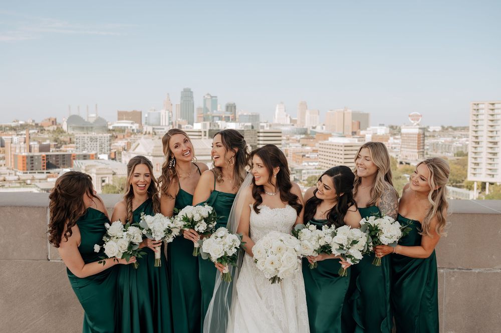 Bridesmaids in emerald green gowns holding white bouquets laugh with bride on a city rooftop.