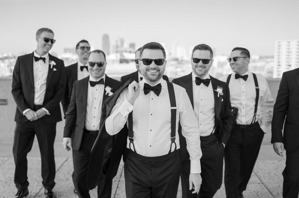 Groom and groomsmen in black tuxedos with sunglasses pose confidently on a rooftop in black and white.