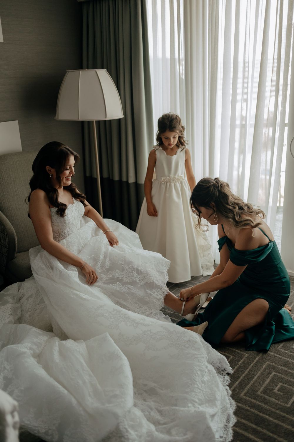 Bride in a white gown getting shoes put on by bridesmaid in emerald dress with flower girl.