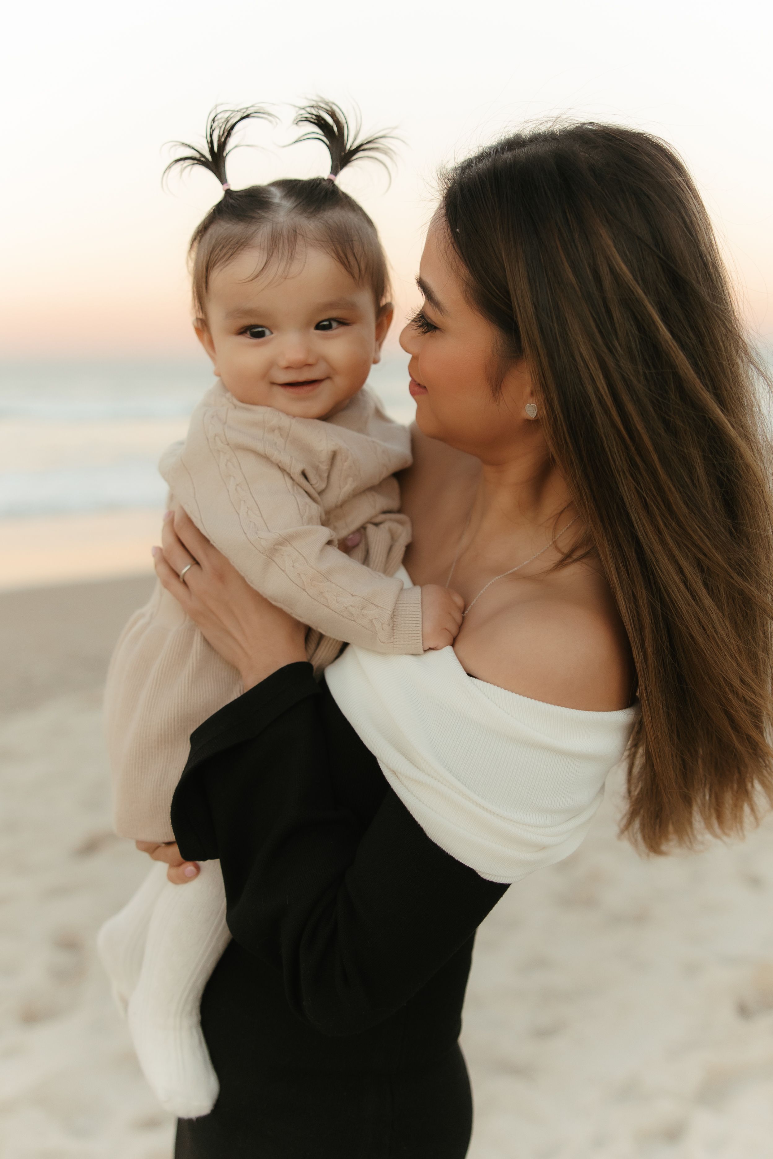 A tender moment captured between parent and baby on a sandy beach at sunset in matching white outfits.