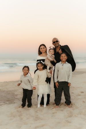 A family wearing coordinated white and black outfits poses together on a sandy beach at sunset.