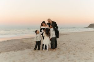 Family group wearing white outfits poses together on sandy beach at sunset.