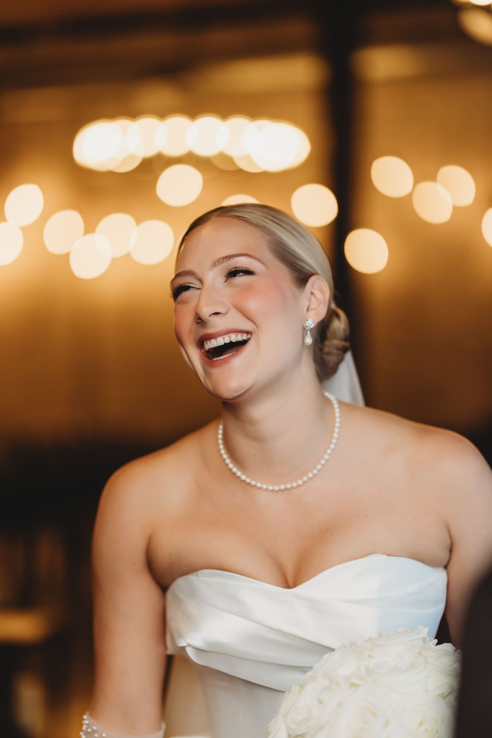 Bride wearing strapless white wedding dress laughs joyfully with pearl necklace against warm bokeh background.