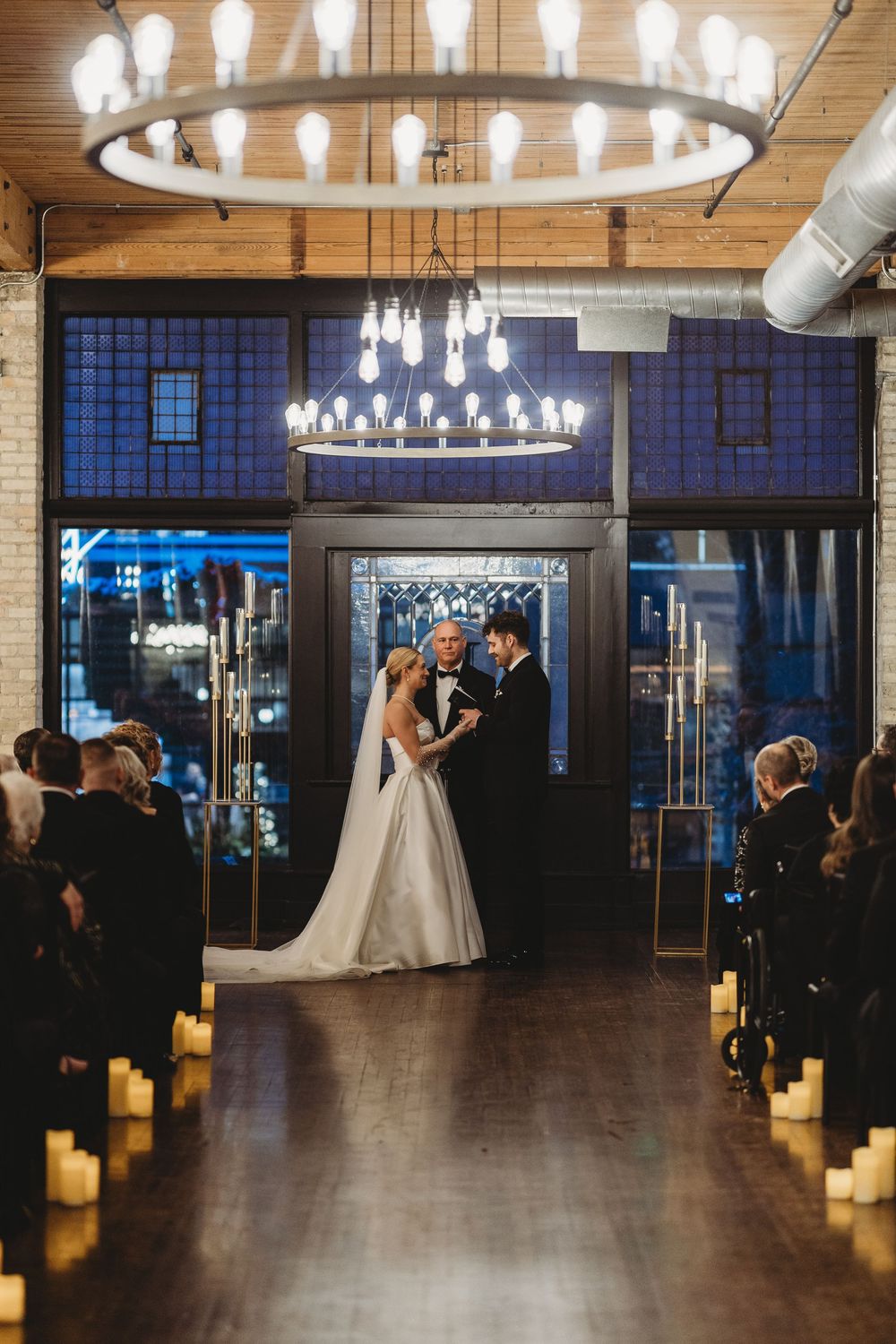 Elegant wedding venue with circular chandeliers and candlelit aisle lined with glowing pillar candles on a dark wood floor.