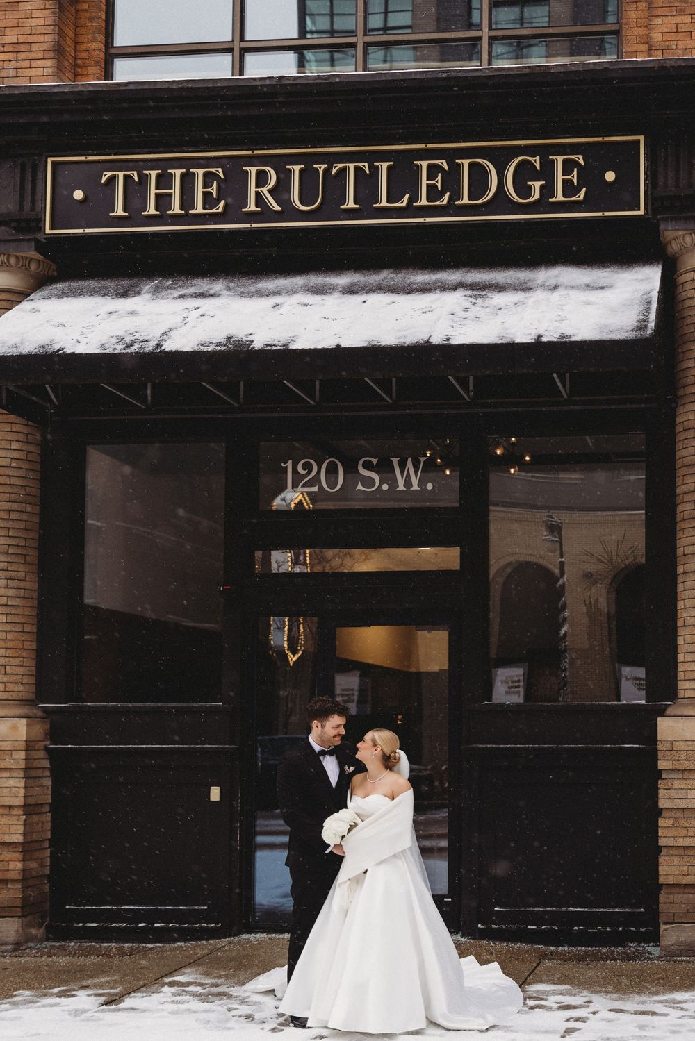 A couple in wedding attire poses in front of The Rutledge venue storefront with a snowy winter backdrop.