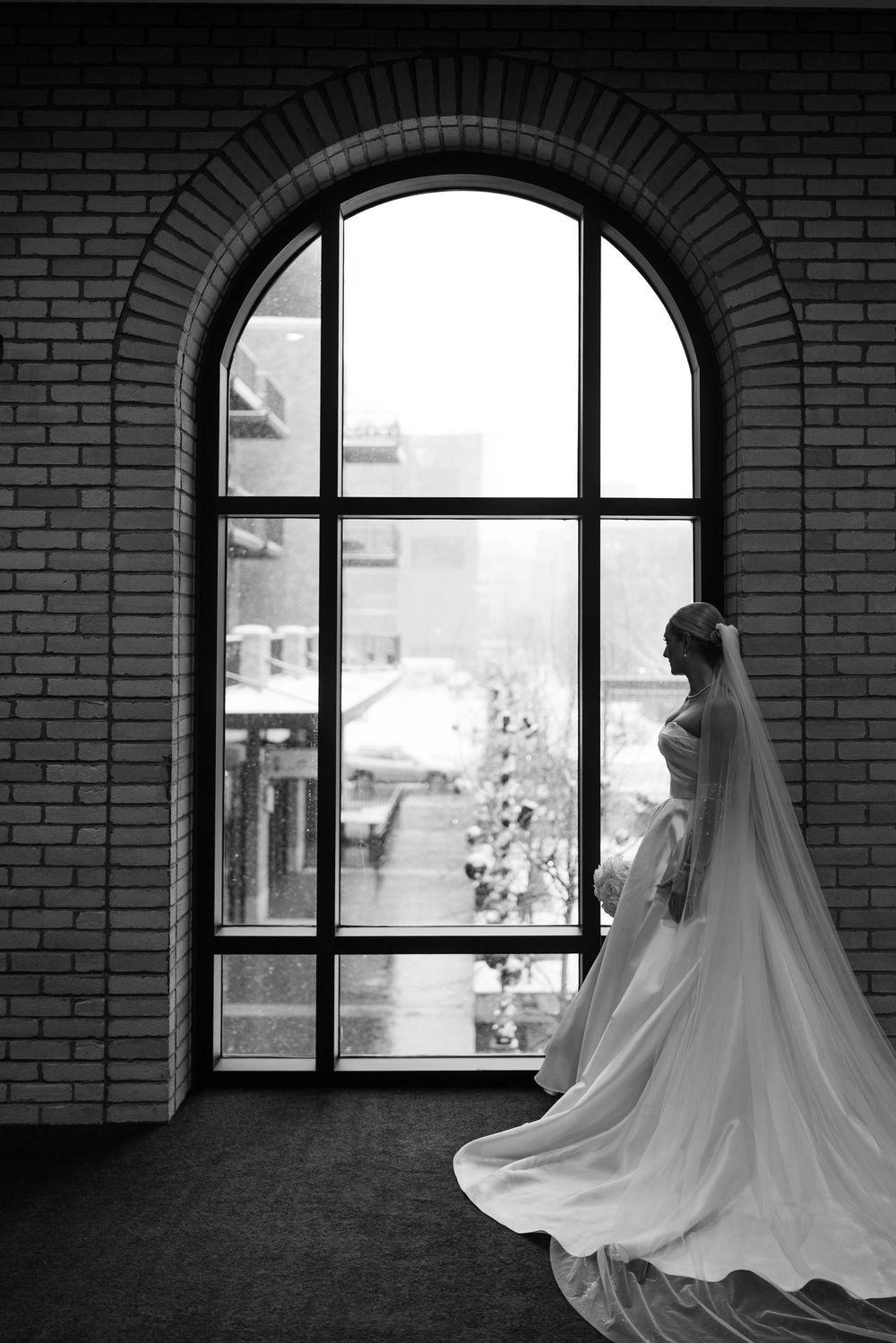 Silhouette of bride in flowing gown and veil standing by arched window in dramatic black and white photo.