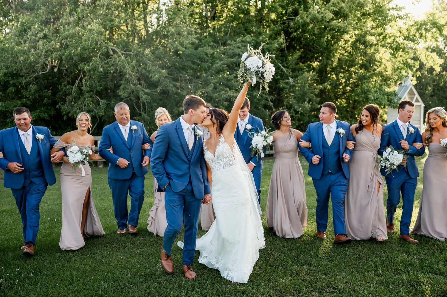 Wedding party poses informally on grass with bridesmaids in neutral dresses and groomsmen in blue suits.