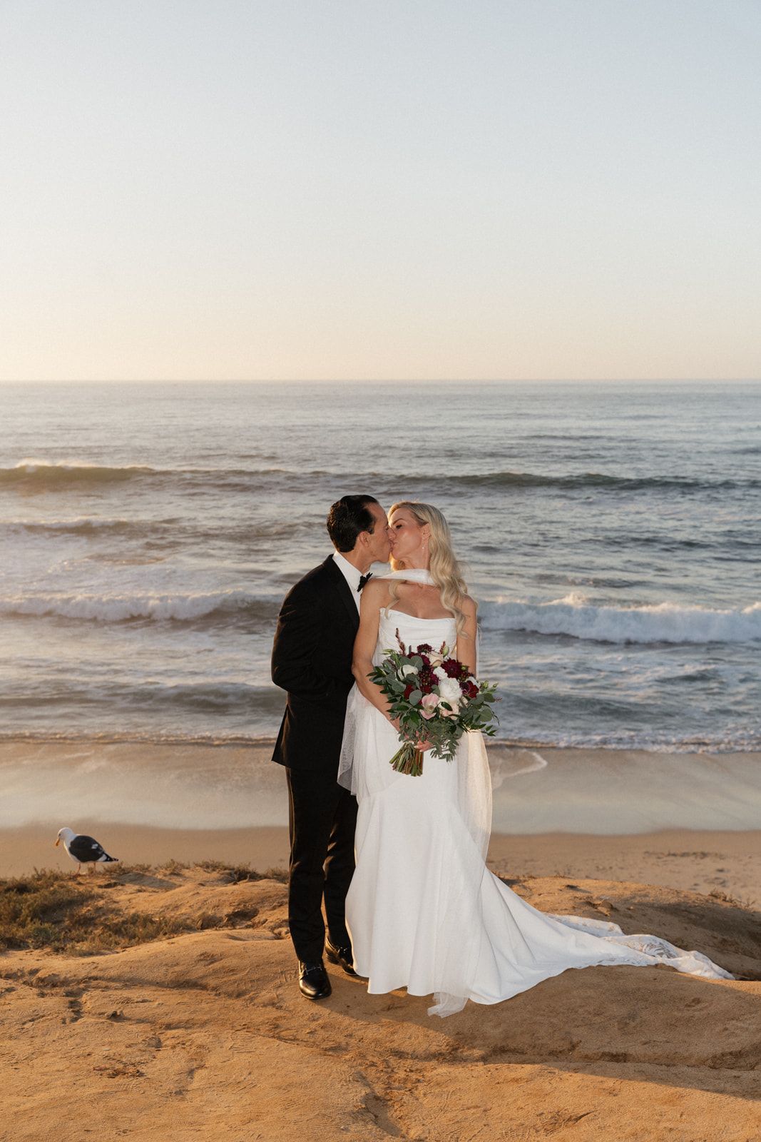 A couple shares a romantic moment at sunset on a beach with ocean waves in the background.