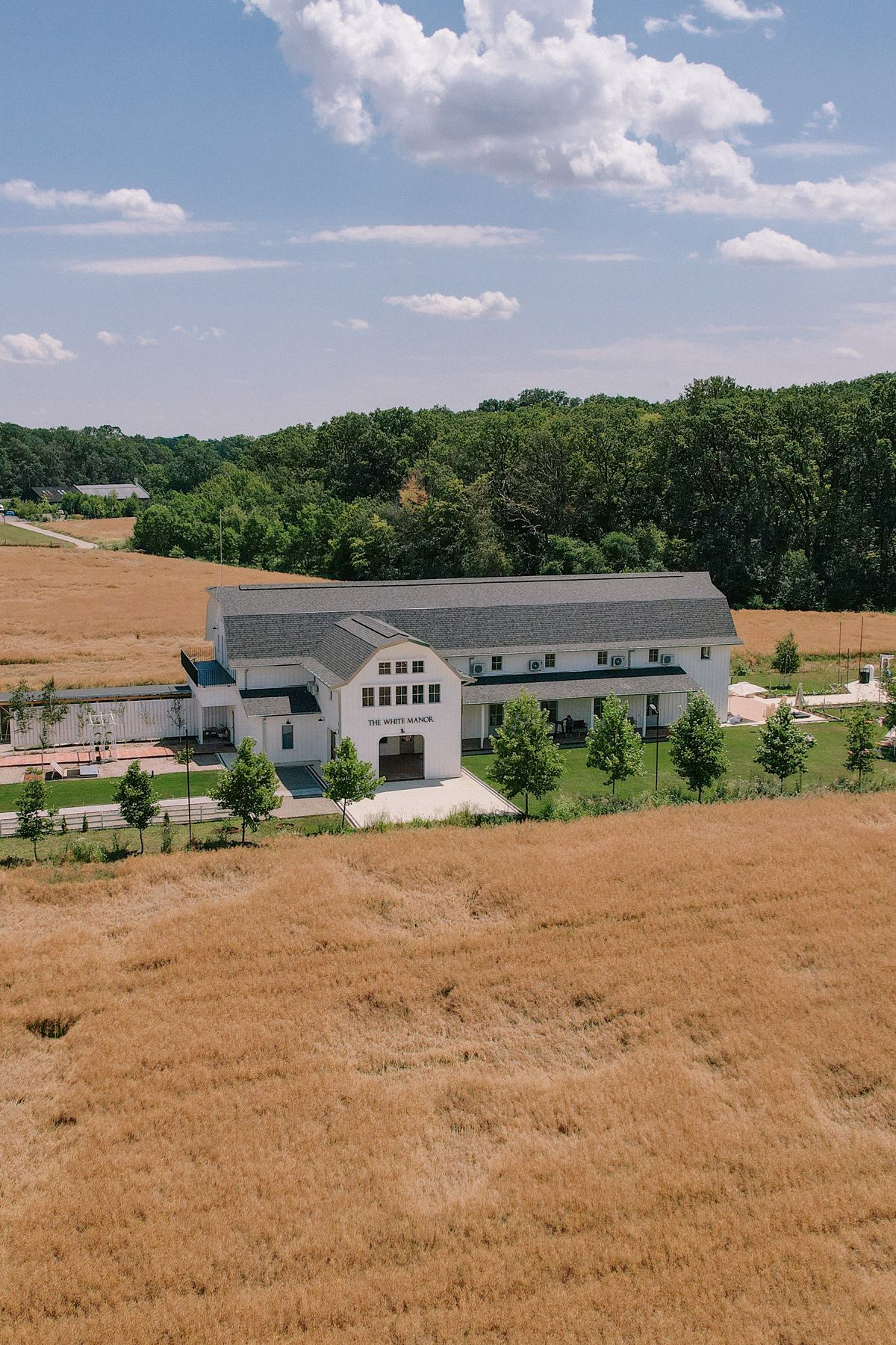 Modern white barn venue surrounded by golden wheat fields and green trees on a sunny summer day.