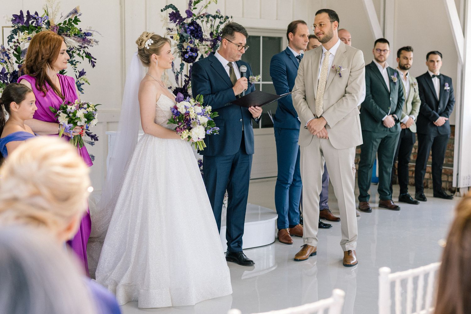 Wedding ceremony taking place in a bright white chapel with purple floral accents.