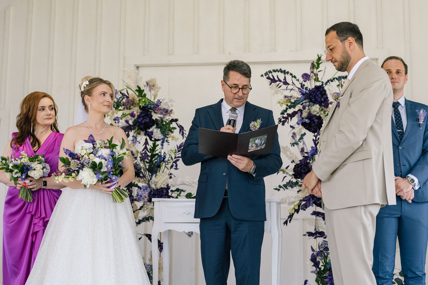 Wedding officiant reads from book during ceremony with bridal party looking on.