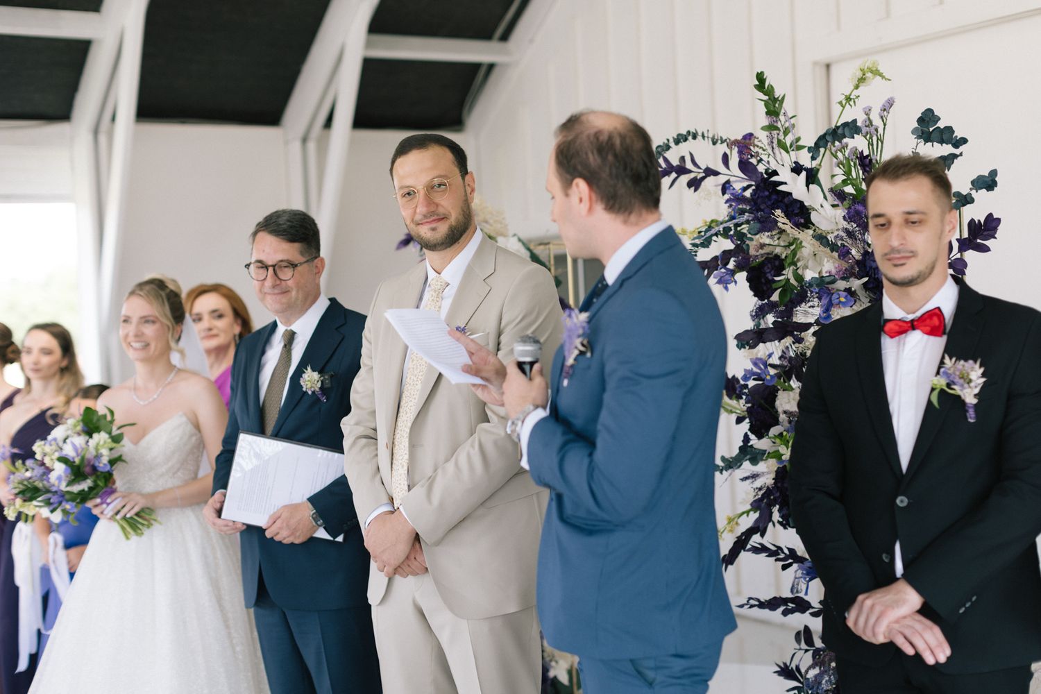Wedding party members in navy and purple dresses gather during an indoor ceremony in a bright white venue.