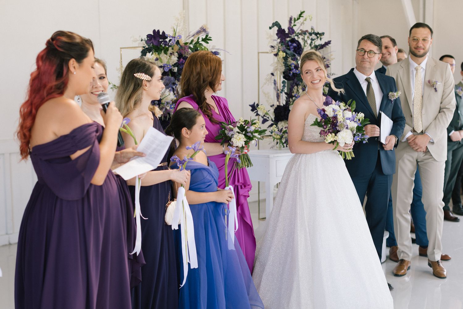 Bridesmaids in coordinating purple and blue dresses stand together during a wedding celebration.