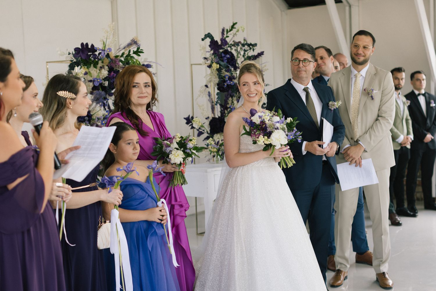 Wedding ceremony participants stand in formal attire against white paneled walls.
