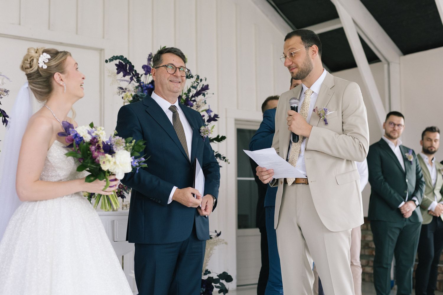 Wedding guests and party members mingle in a bright white indoor venue with vertical wall paneling.