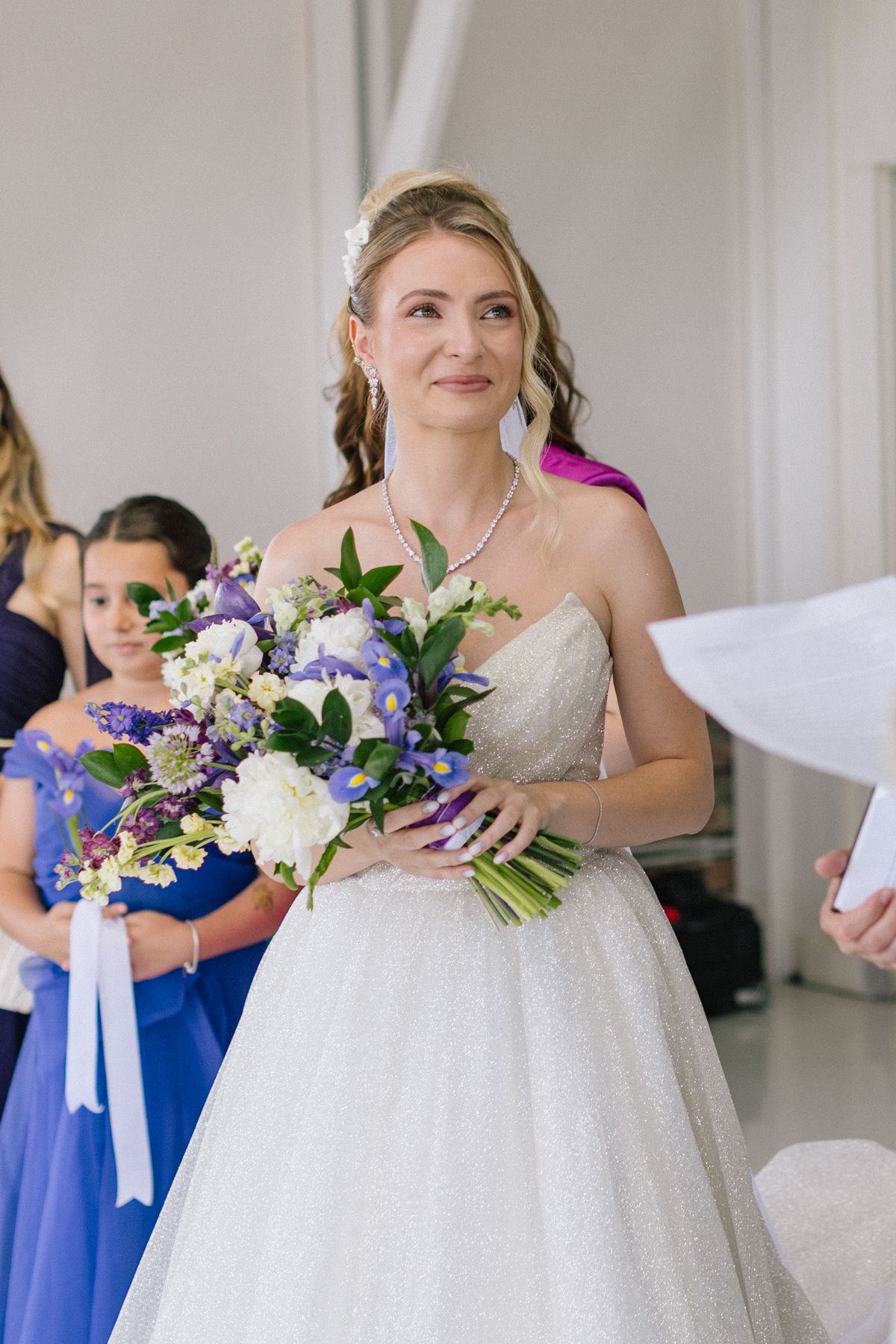 Wedding party members hold beautiful purple and blue floral bouquets in a bright indoor setting.