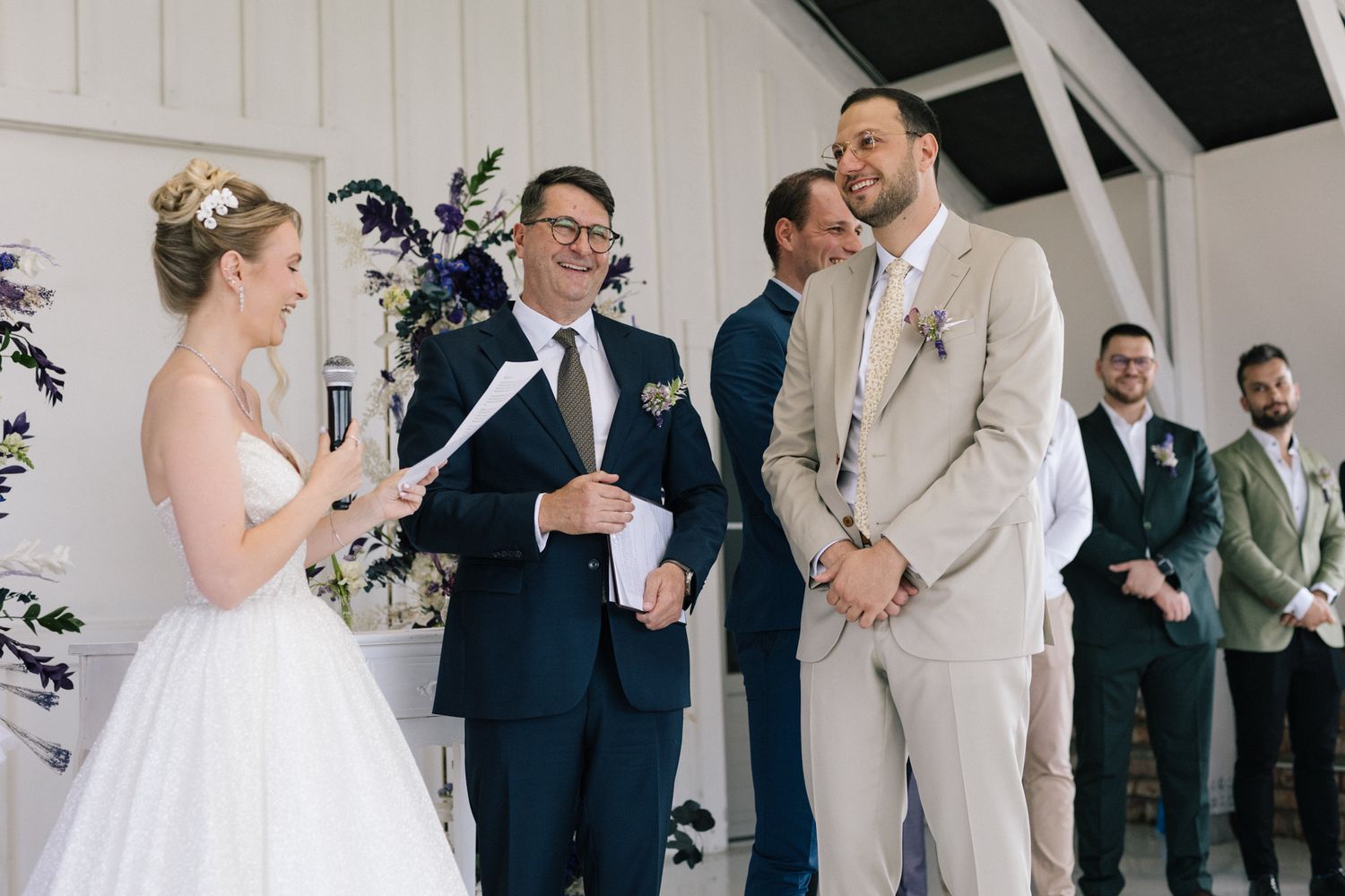 Wedding ceremony participants stand in formal attire inside a bright white venue.