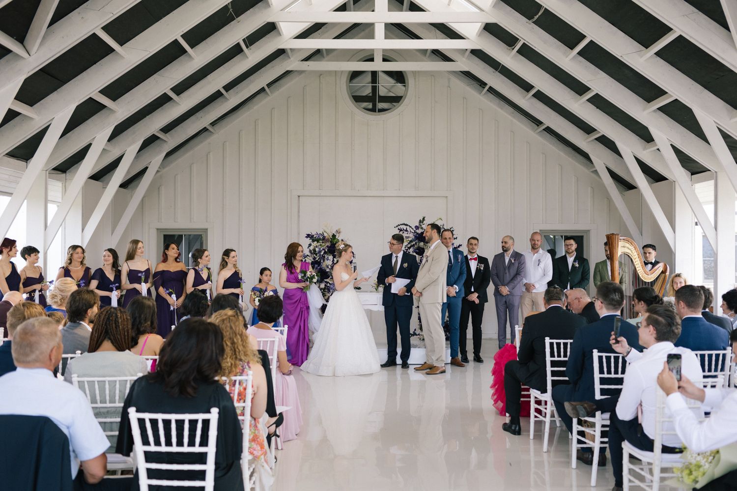 Wedding guests mingle in an elegant white barn venue with exposed beams and white chairs.