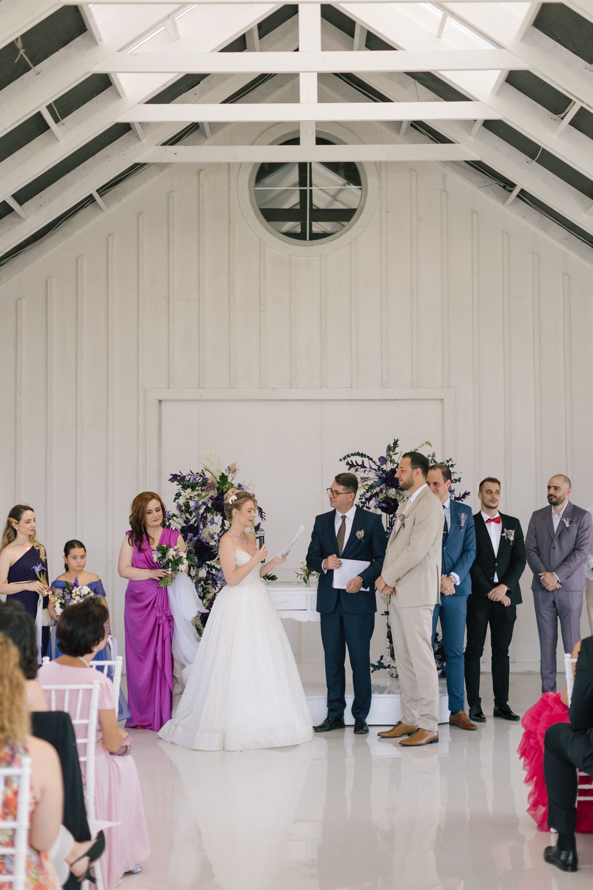 Wide view of wedding ceremony in a white vaulted ceiling venue with guests gathered around.