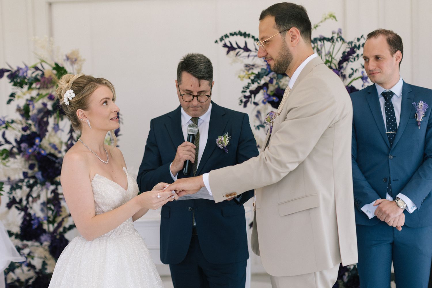 Wedding ceremony proceedings in a light-filled venue with purple and white floral arrangements.