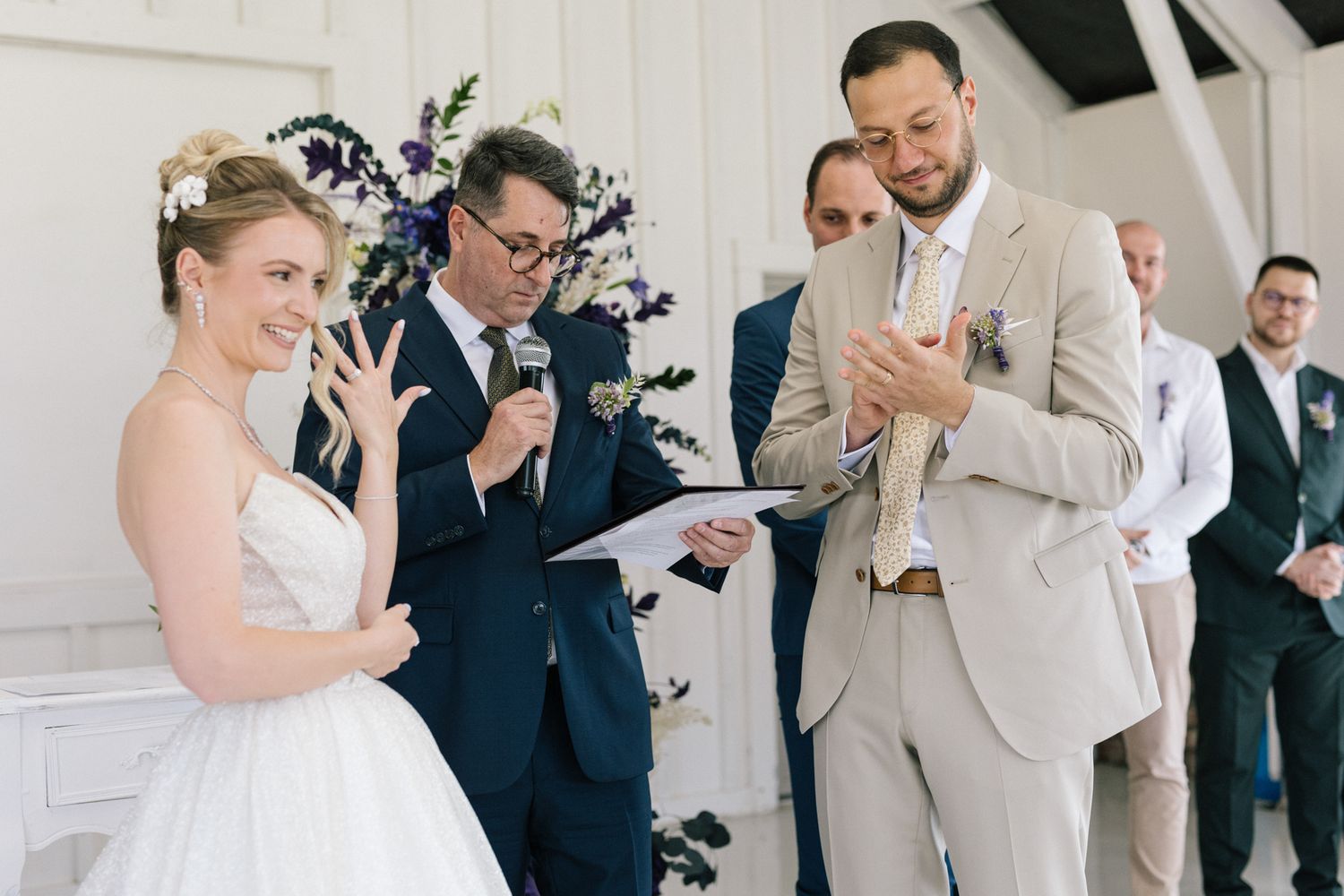 Candid moments during wedding ceremony in a white-walled venue with purple floral accents.