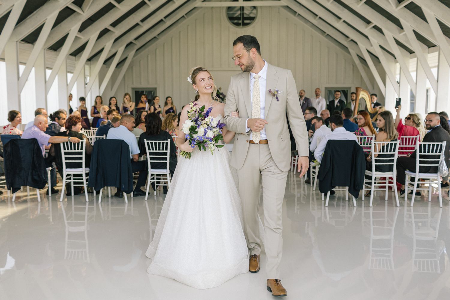 Wedding couple dances together in an elegant white reception venue with exposed beam ceiling and guests seated at tables.