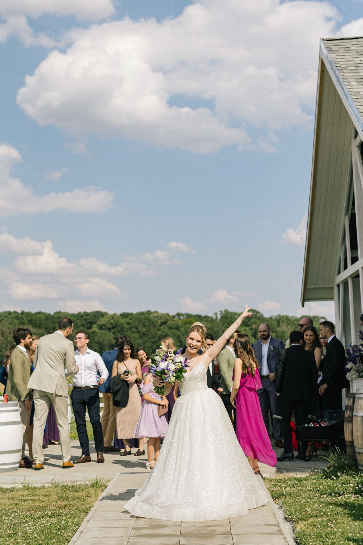 Wedding party and guests gather outside a white building on a sunny day with blue skies.