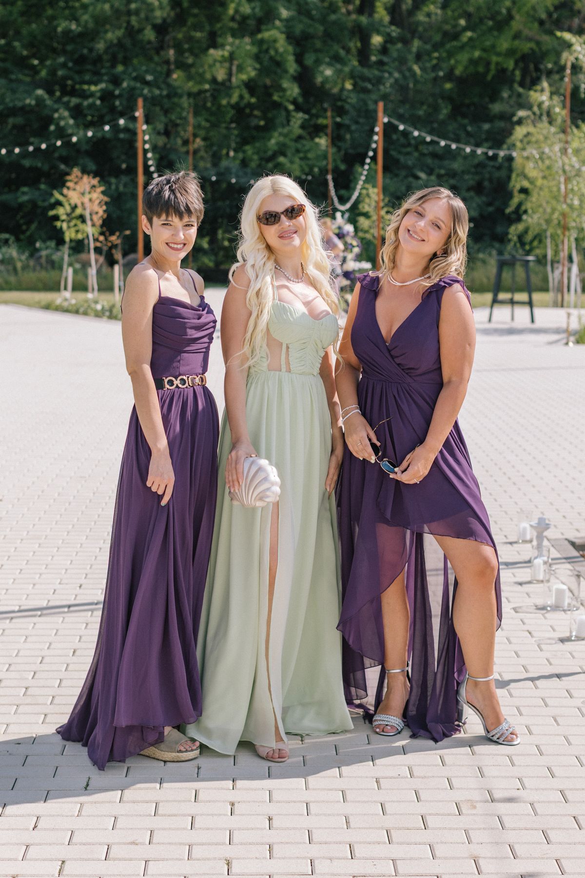 Bridesmaids in elegant purple and sage green dresses pose together on a stone patio at an outdoor wedding venue.