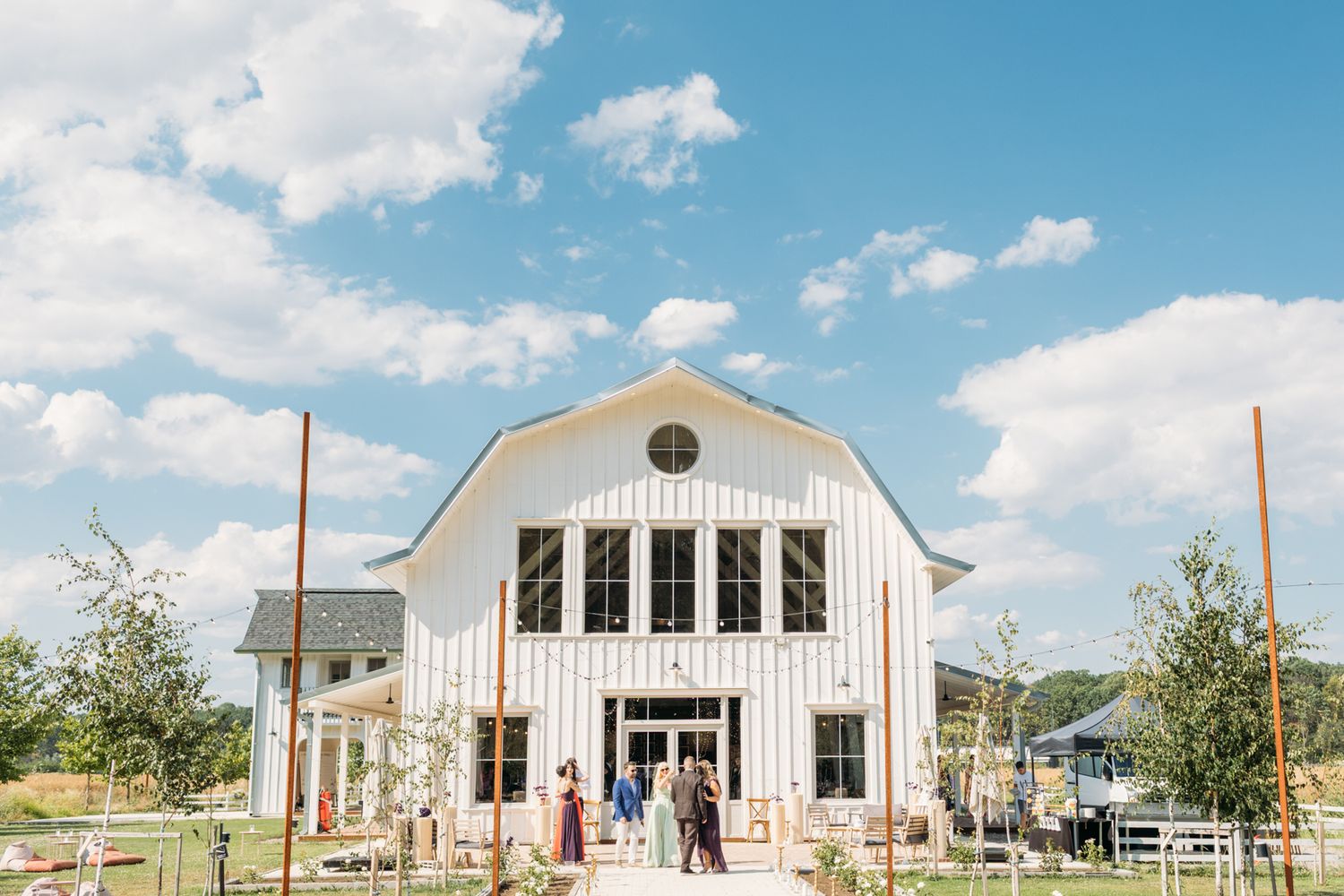 Modern white barn wedding venue with large windows and string lights under a bright blue summer sky.