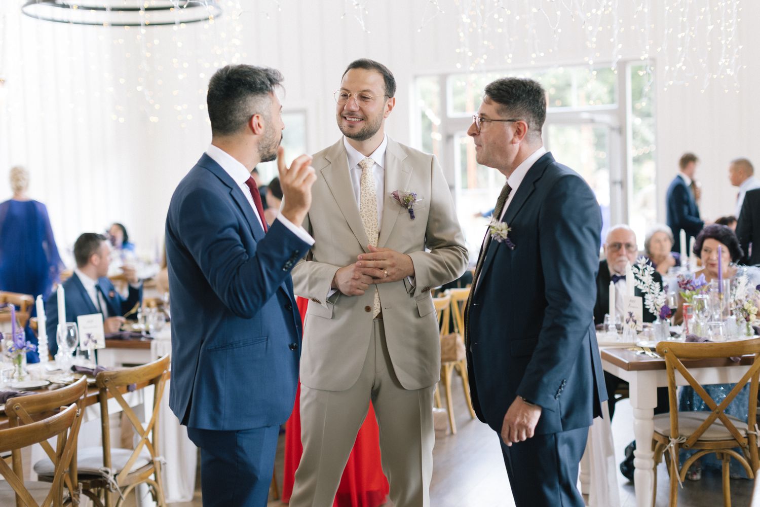 Wedding reception guests mingle and socialize in an airy white venue with wooden cross-back chairs.