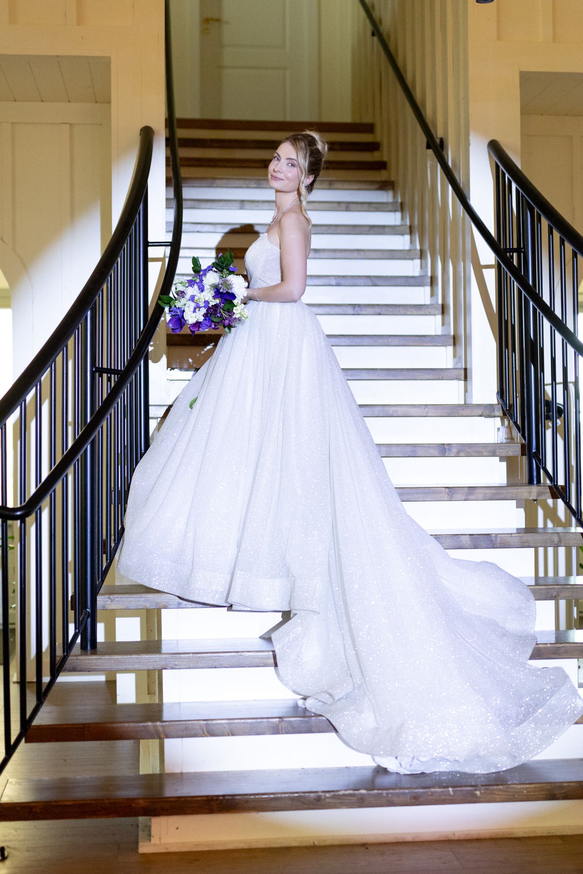 Wedding dress train cascades down elegant wooden staircase with black railings in natural light.