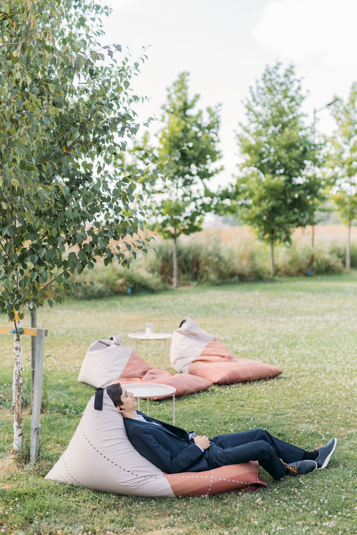 Wedding party members lay on blankets in a grassy field near young trees for creative photography.