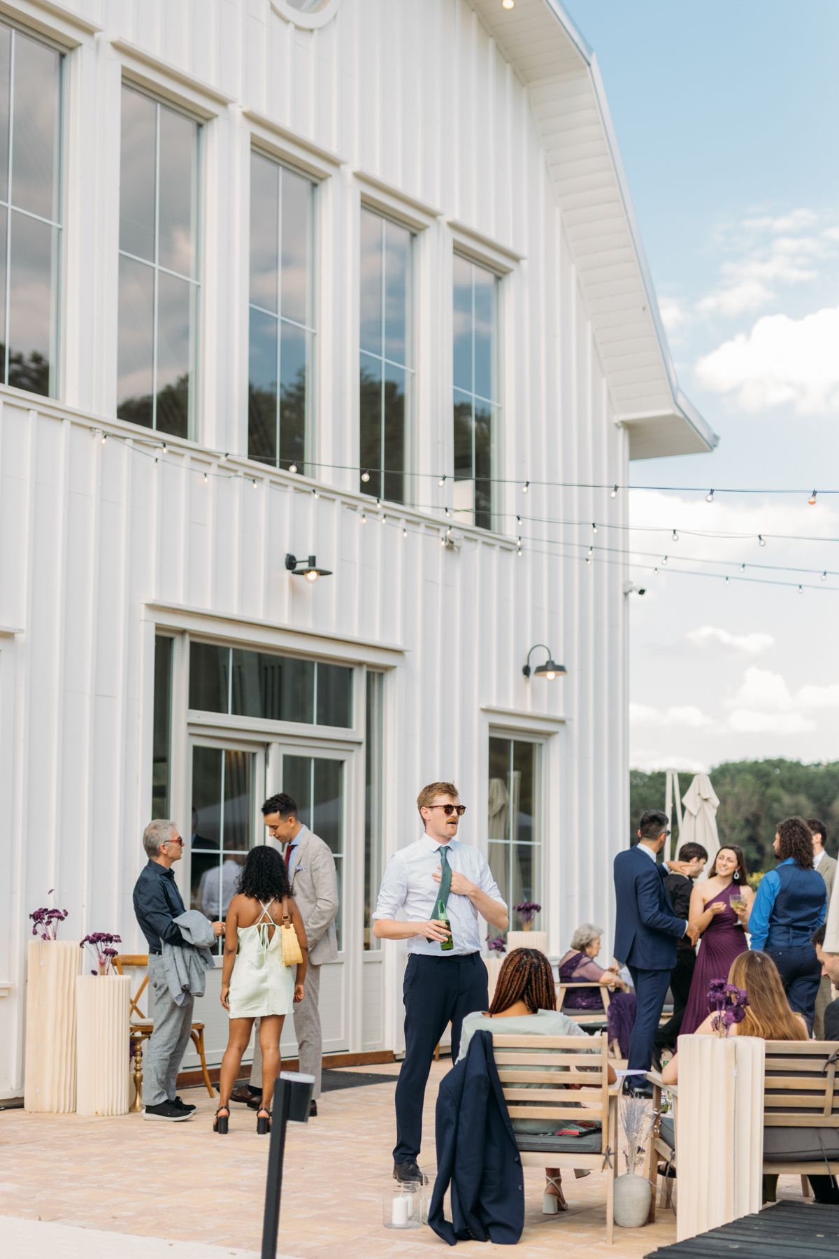Guests gather outside a modern white barn venue with large windows during a wedding reception.
