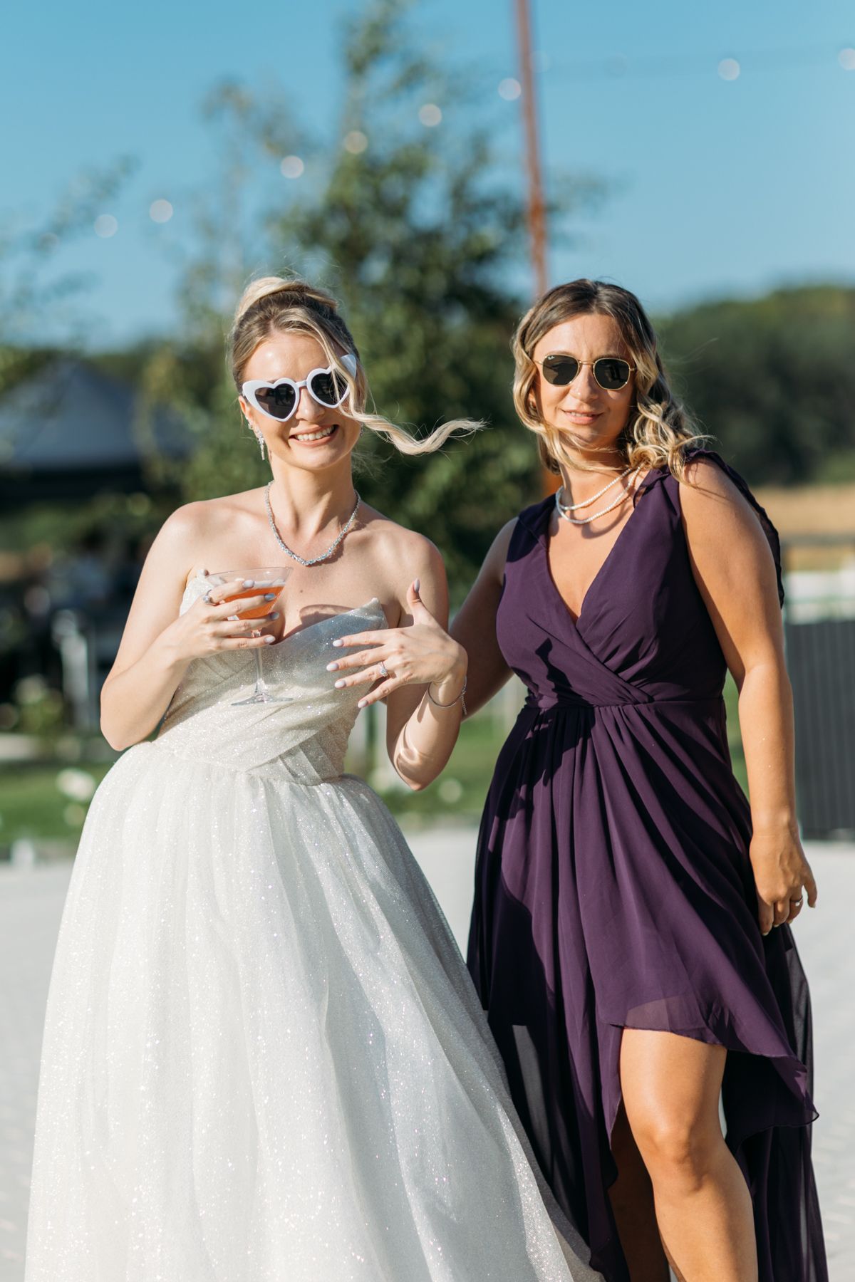 Two women in sunglasses pose together, one wearing a white wedding gown and one in a purple dress.