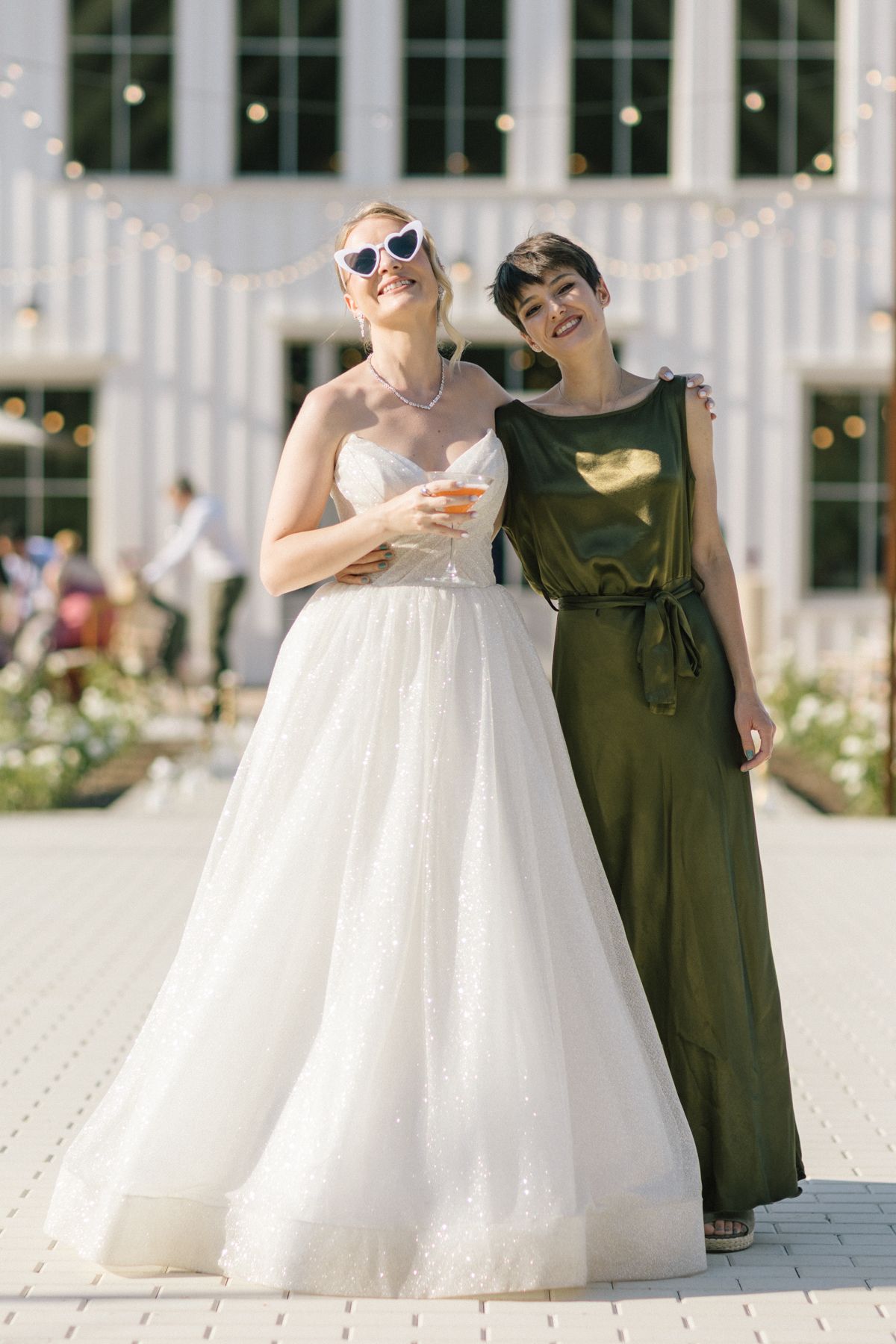 Two women pose together outdoors, one in a white wedding gown and one in an olive green dress.