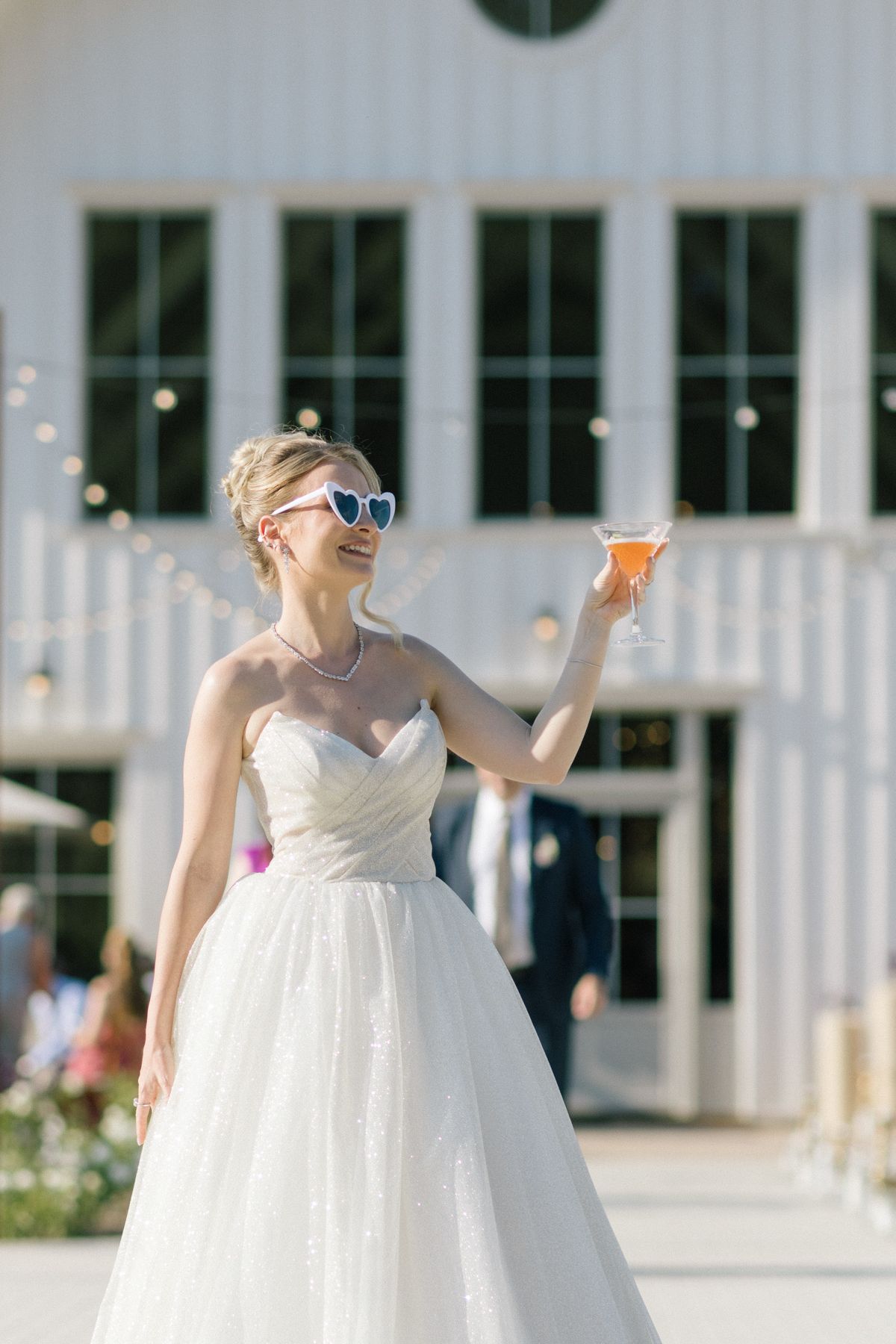 Woman in a white wedding gown poses playfully with a drink outdoors.