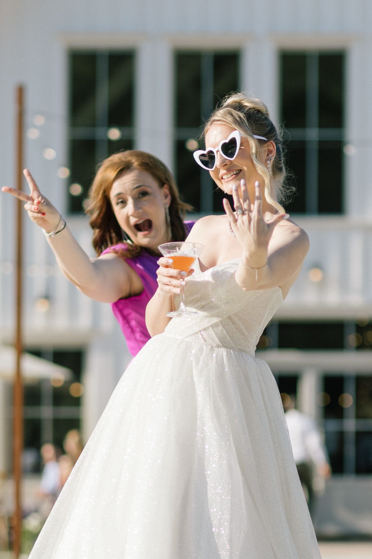 Two women celebrate joyfully outdoors, one in a white wedding gown wearing heart sunglasses.