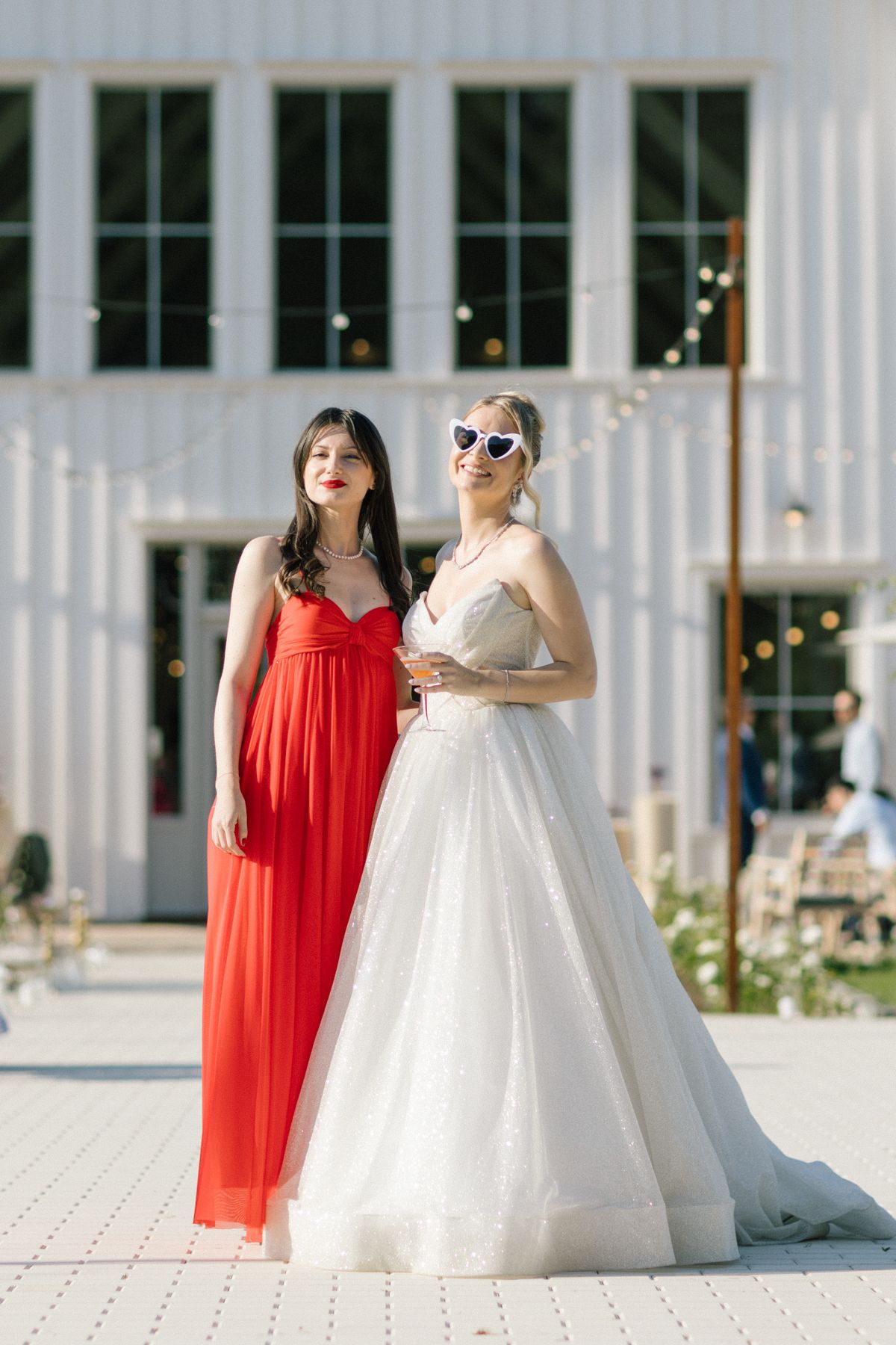 Two women pose together, one in a red dress and one in a white wedding gown, in front of a white building.
