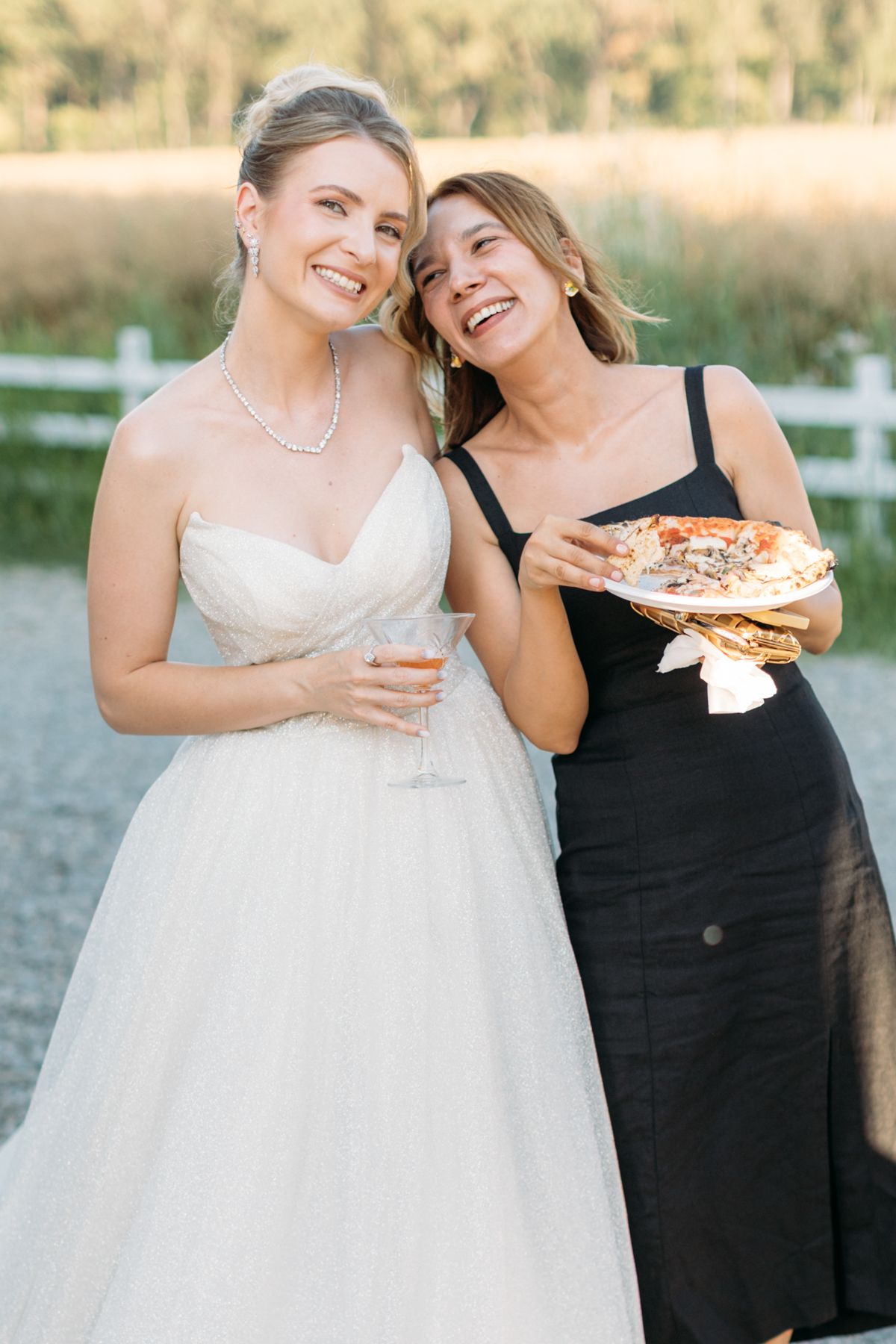 Two women share pizza and laugh together, one in a wedding dress and one in black.