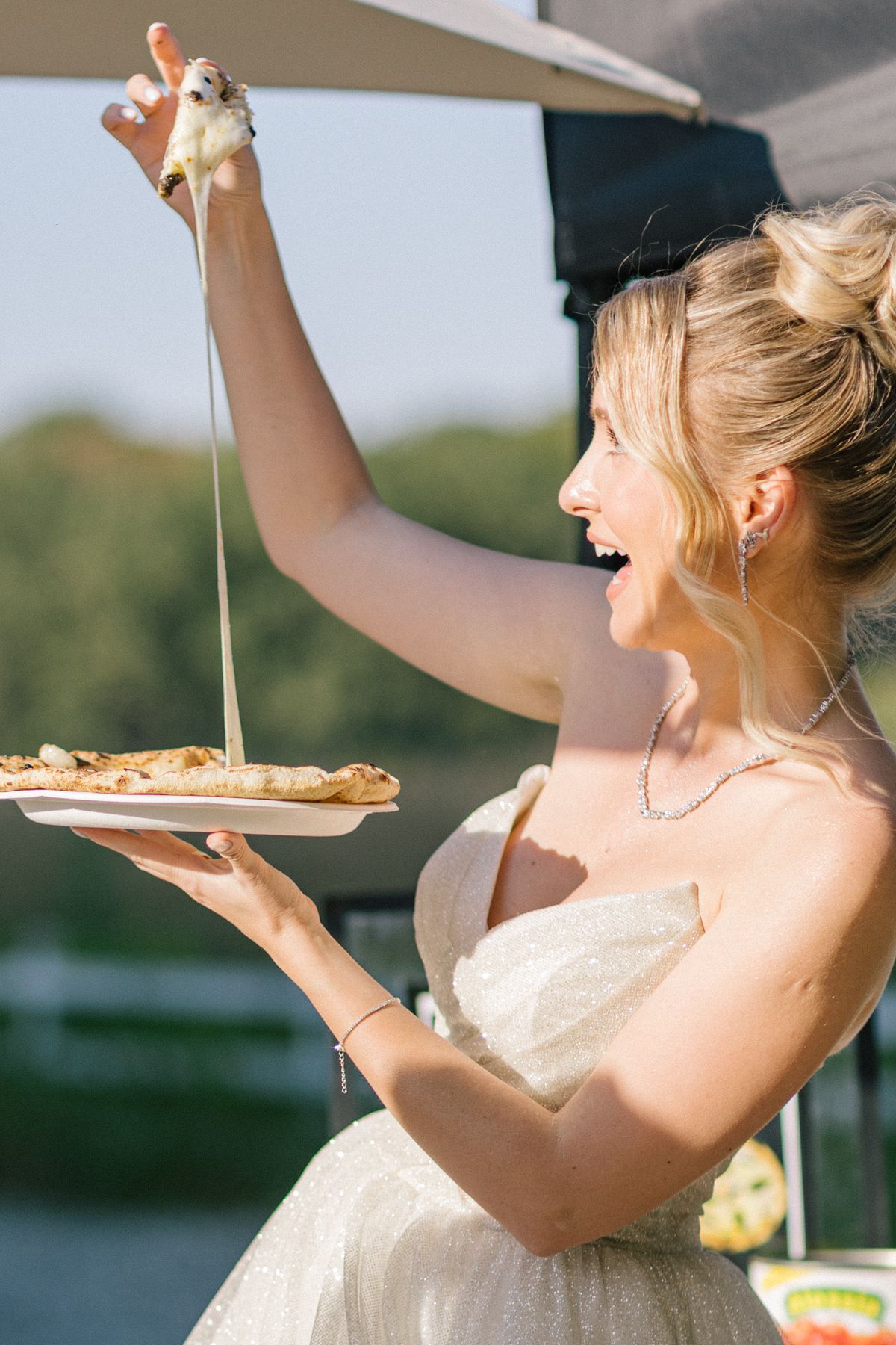 Woman in wedding dress joyfully reaches for pizza at an outdoor venue.