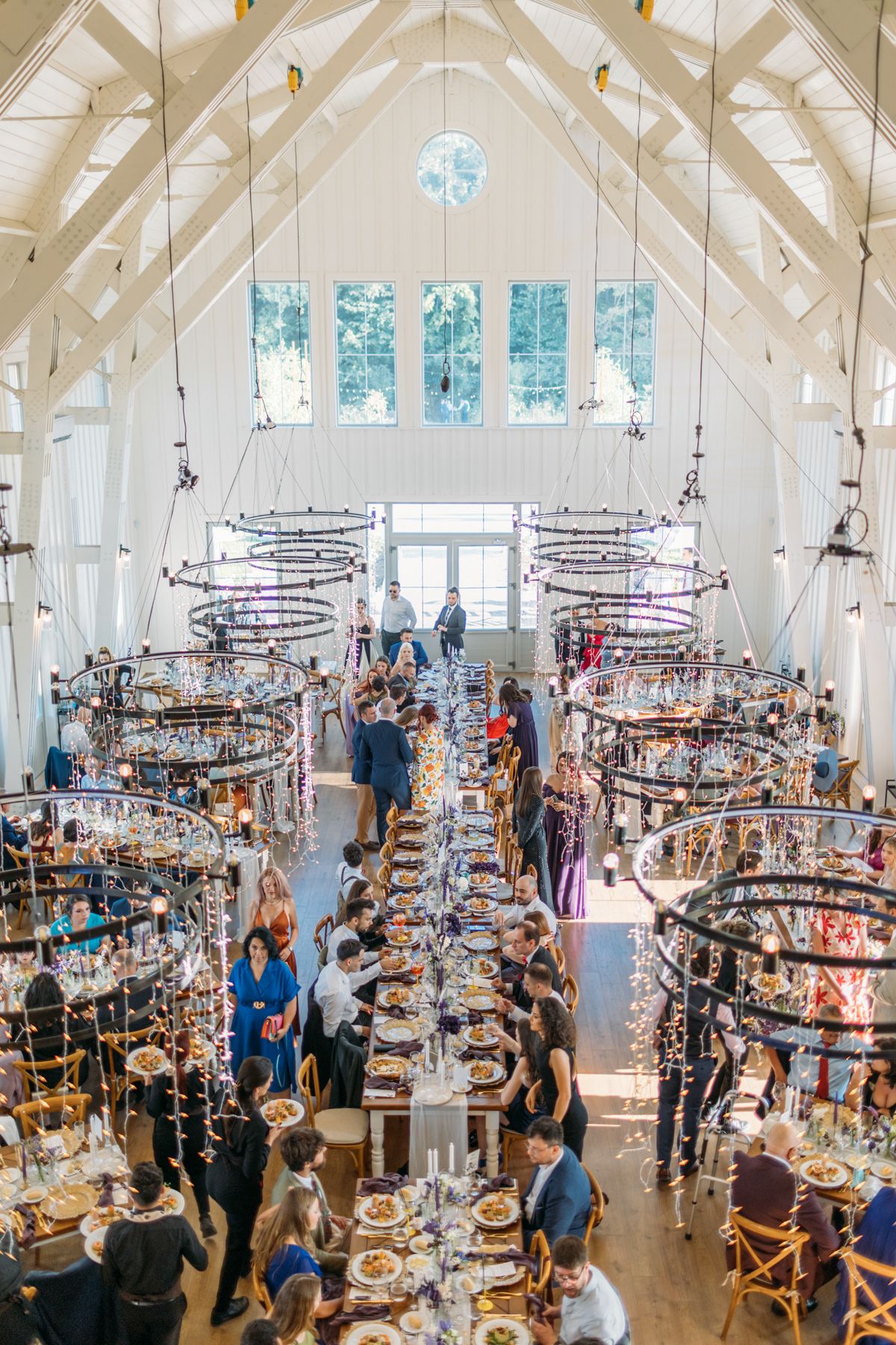 Wedding venue with vaulted white beams and crystal chandeliers filled with guests.