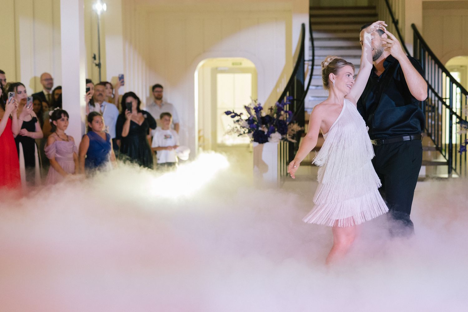 A couple performs their first dance at a wedding reception surrounded by fog effects and guests watching.