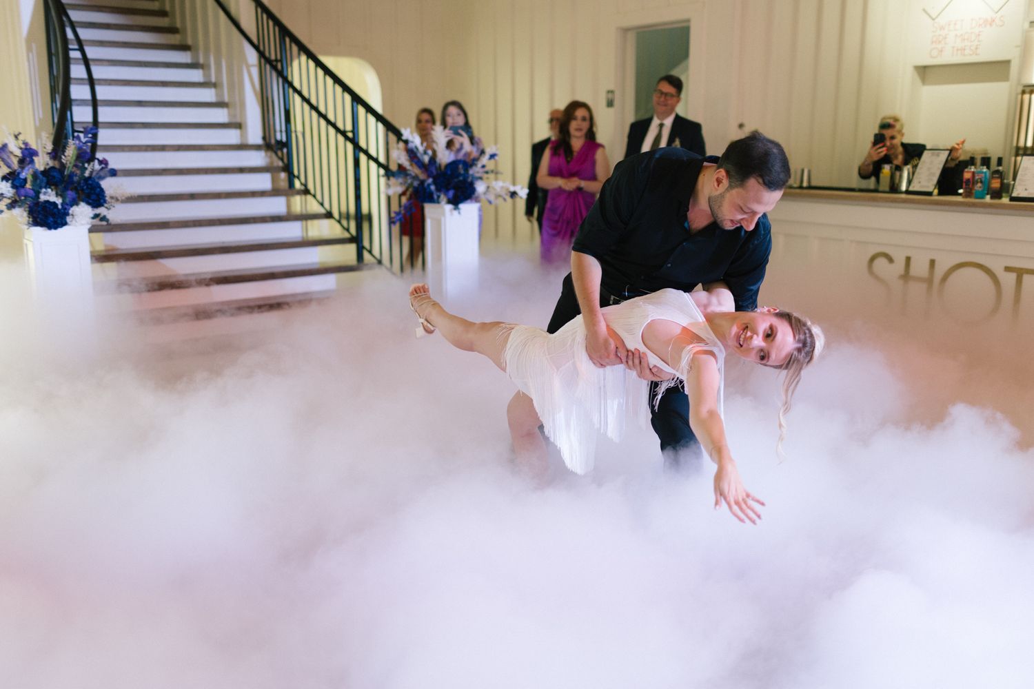 Dancers execute a dramatic dip move on a fog-covered dance floor during a wedding celebration.