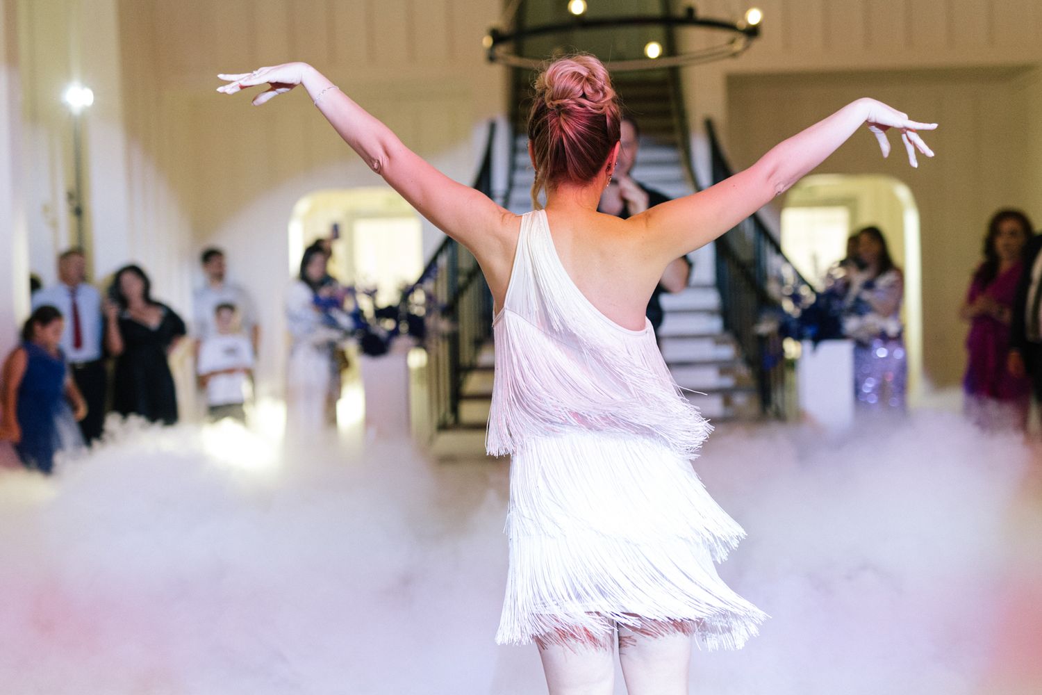 A dancer in a white fringe dress strikes a pose with arms raised amid swirling fog on the dance floor.