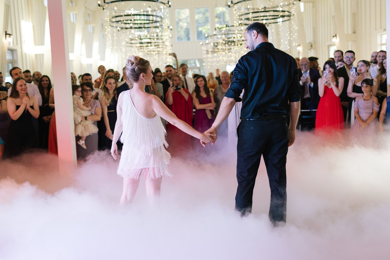 Dancers move gracefully through low-lying fog under elegant chandeliers at a wedding reception.