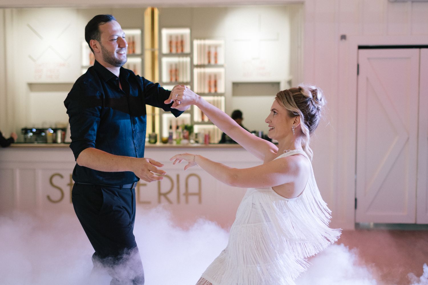 A couple performs an upbeat dance routine in a white-walled venue with fog effects.