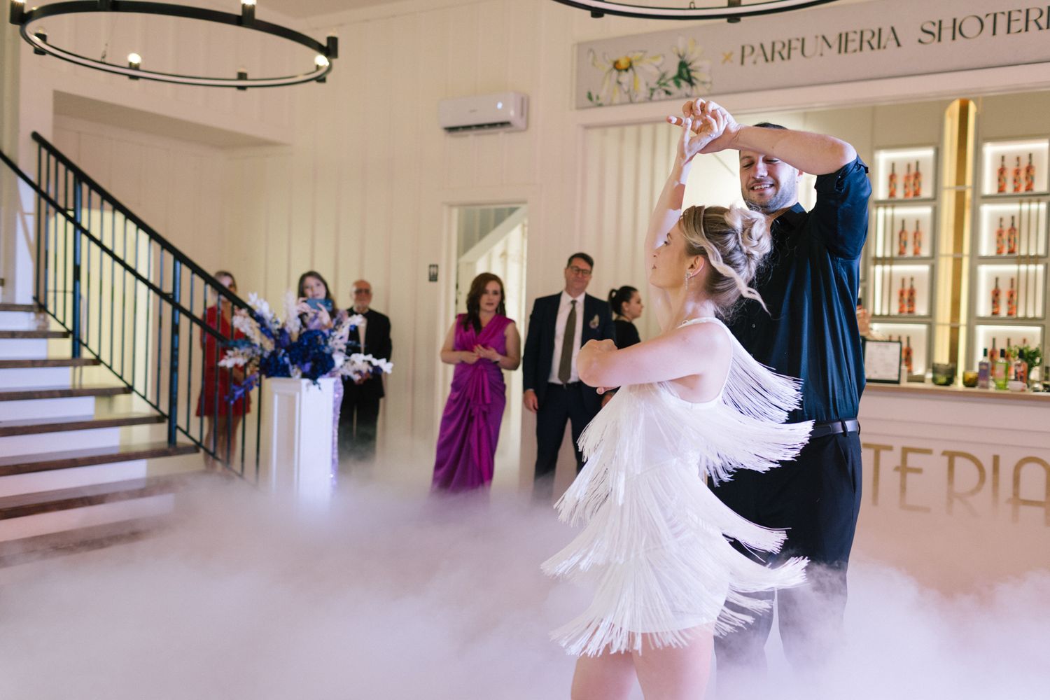 Wedding dancers perform under a circular chandelier with fog effects and guests watching from the staircase.