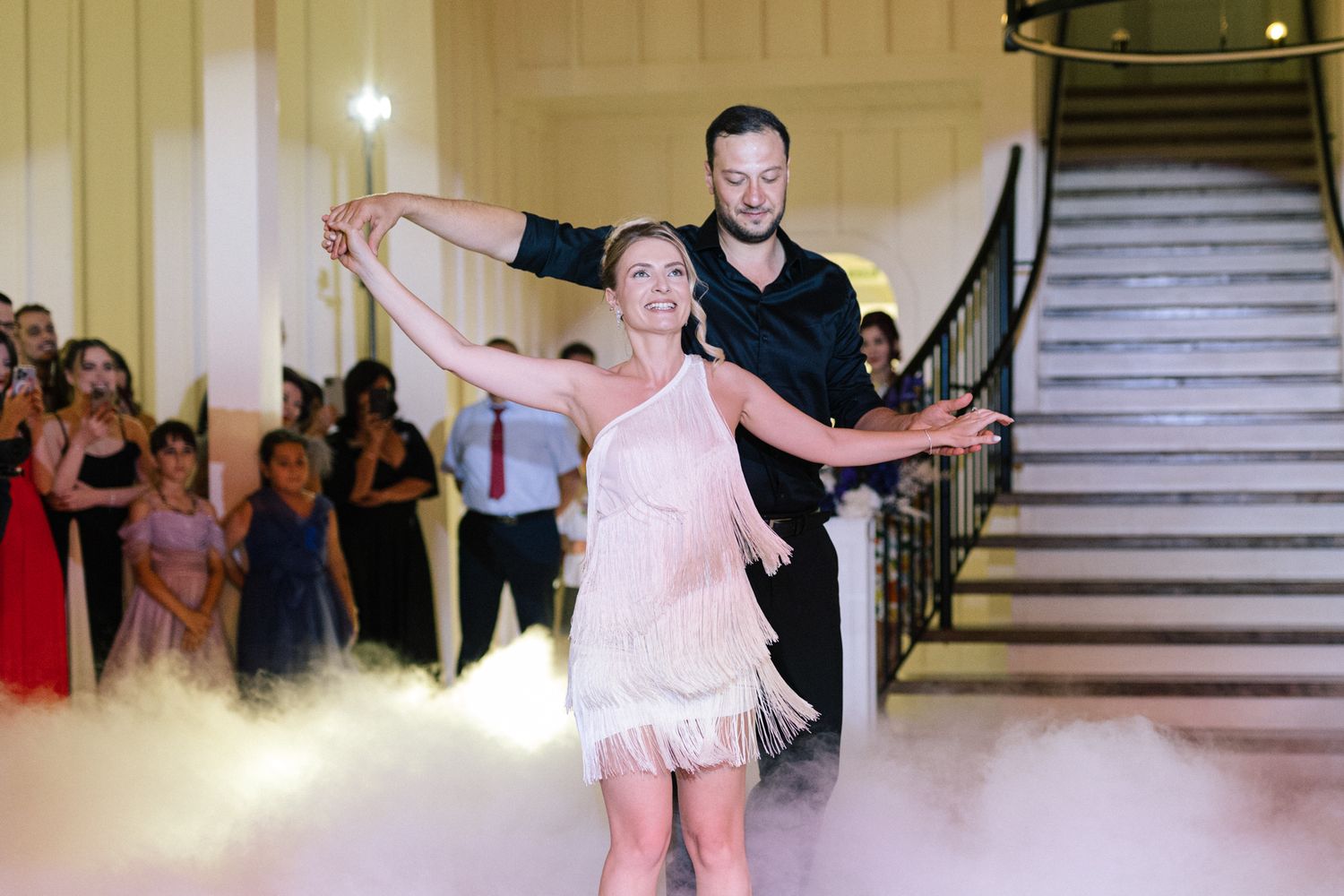 A dance performance unfolds on a fog-filled floor with dramatic lighting and guests watching.