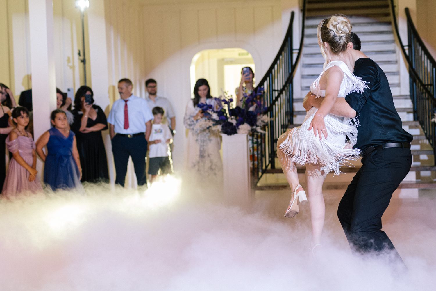 Wedding dancers perform on a fog-covered floor while guests watch from the staircase area.