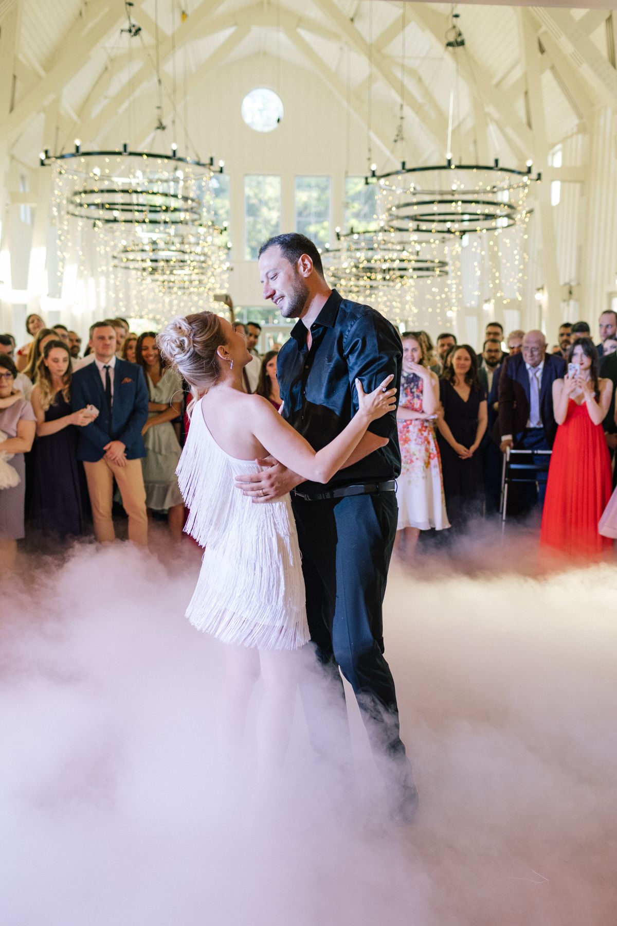 A couple shares their first dance on a misty dance floor under crystal chandeliers at an elegant wedding reception.